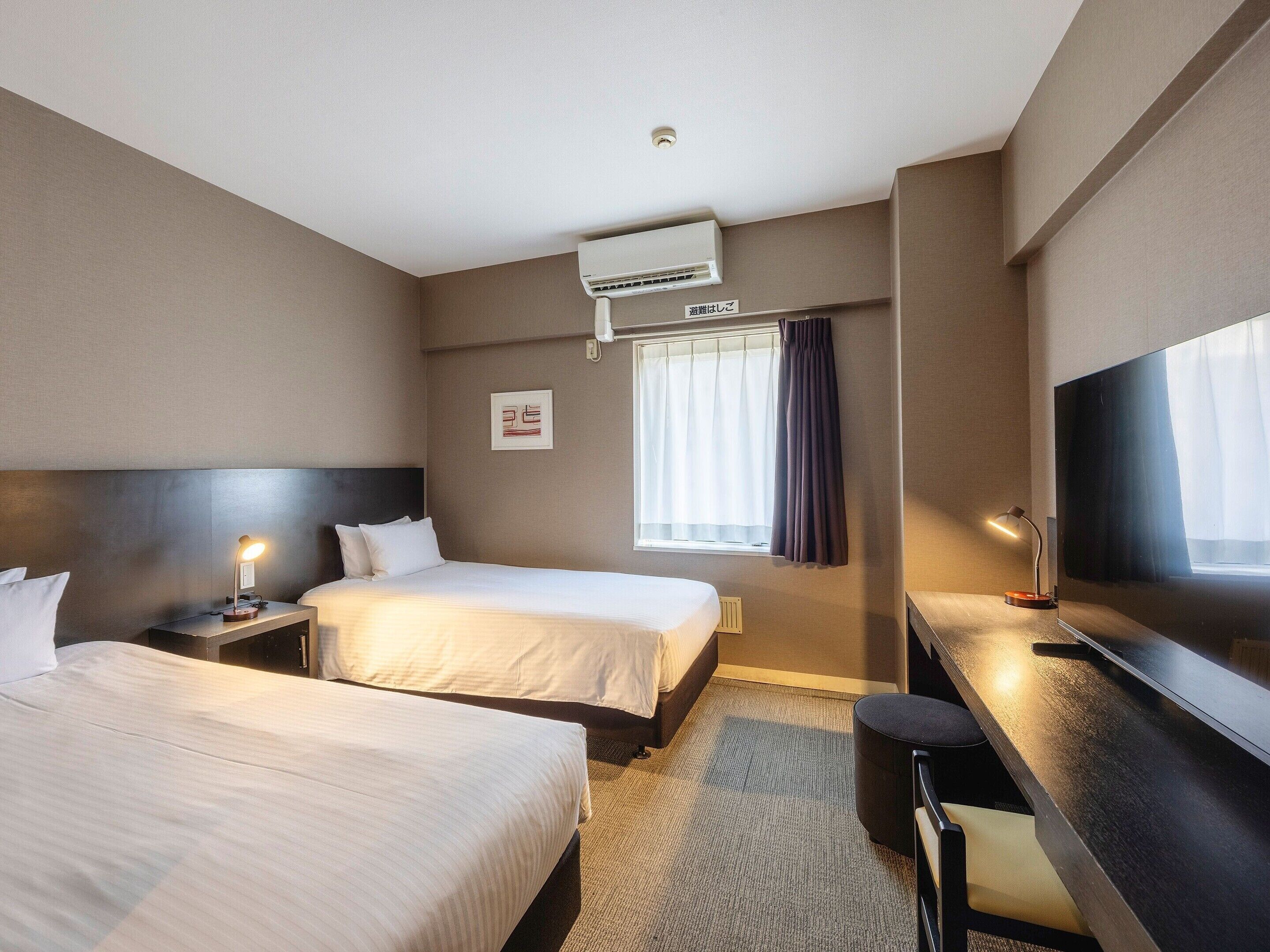 Una habitación de hotel con dos camas, un escritorio, una silla, un equipo de aire acondicionado, una ventana con cortinas y un televisor montado en la pared.