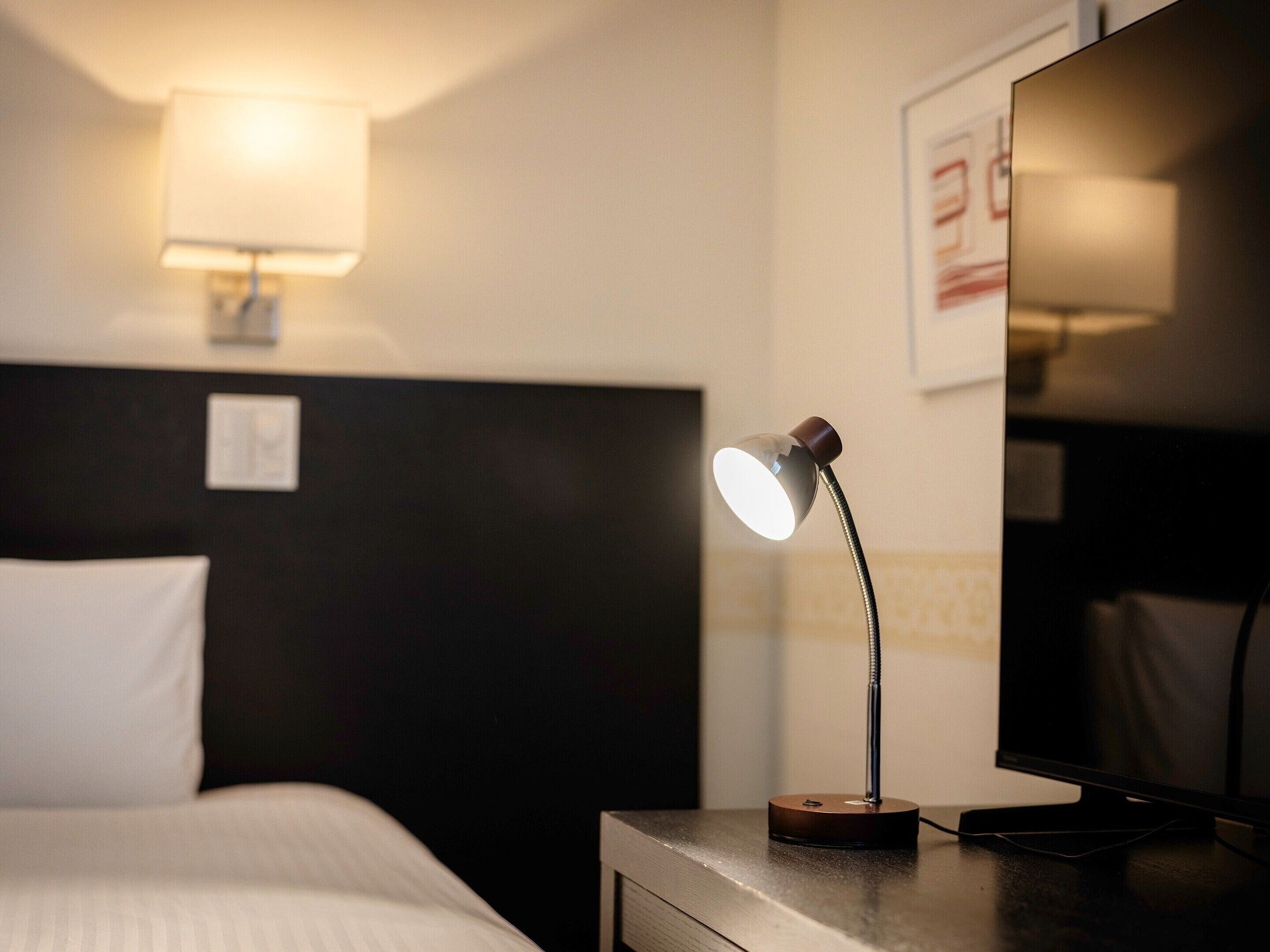 A hotel room with a bed, a desk with a lamp, a flat-screen TV, and a wall-mounted light fixture.