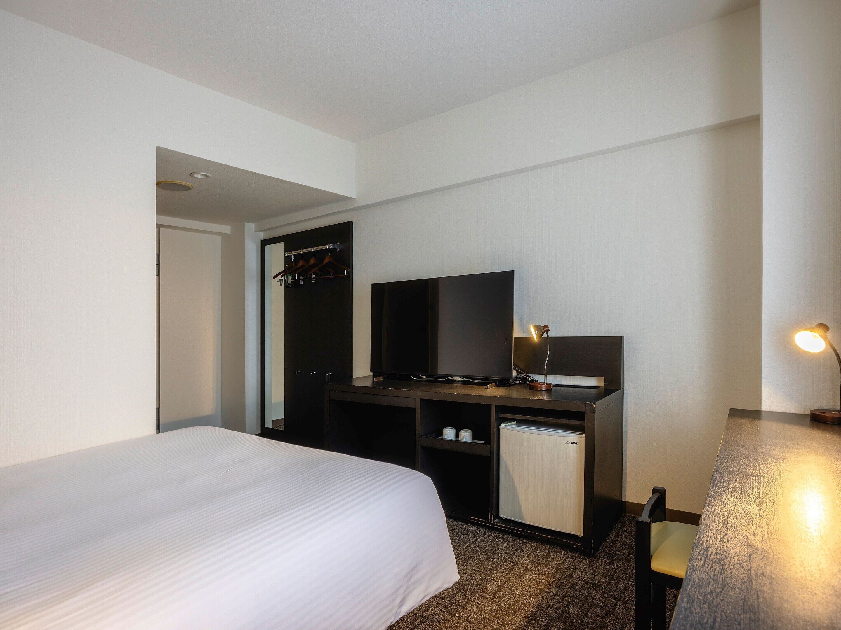 A hotel room with a bed, a desk with a television, a small refrigerator, and a desk lamp.