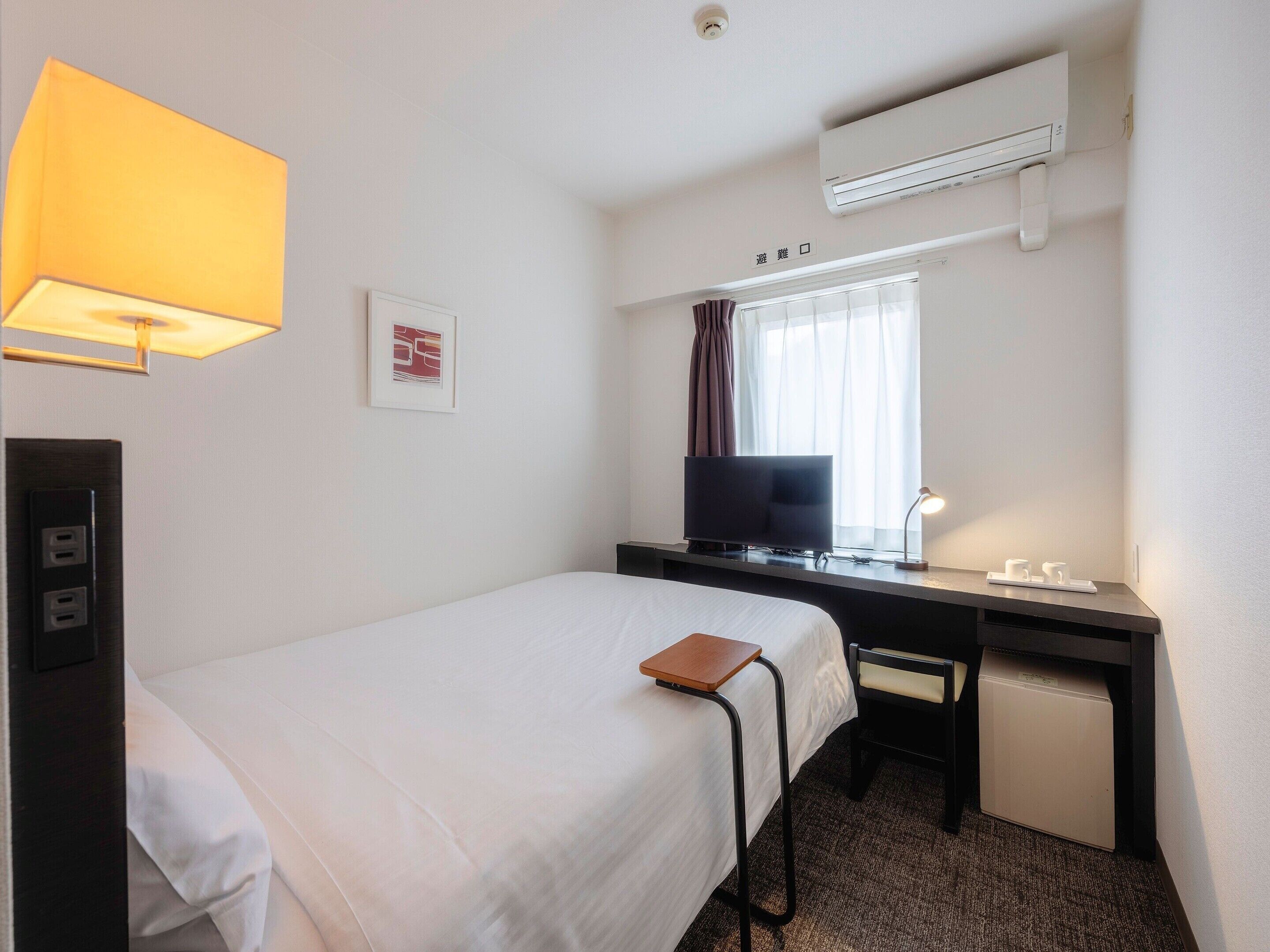 Una habitación de hotel con cama, escritorio, televisor, aire acondicionado y lámpara.