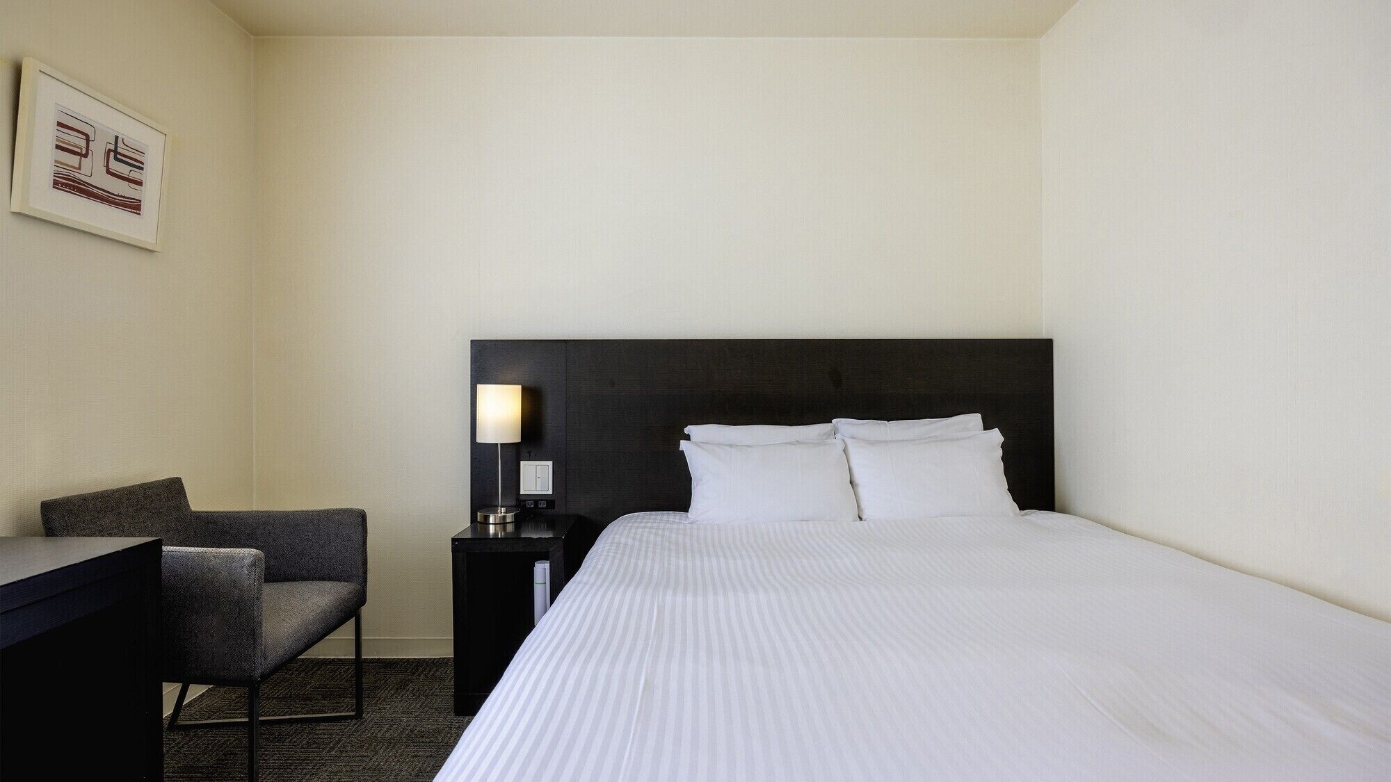 Quarto de hotel com cama, escrivaninha, cadeira e mesa de cabeceira com abajur.