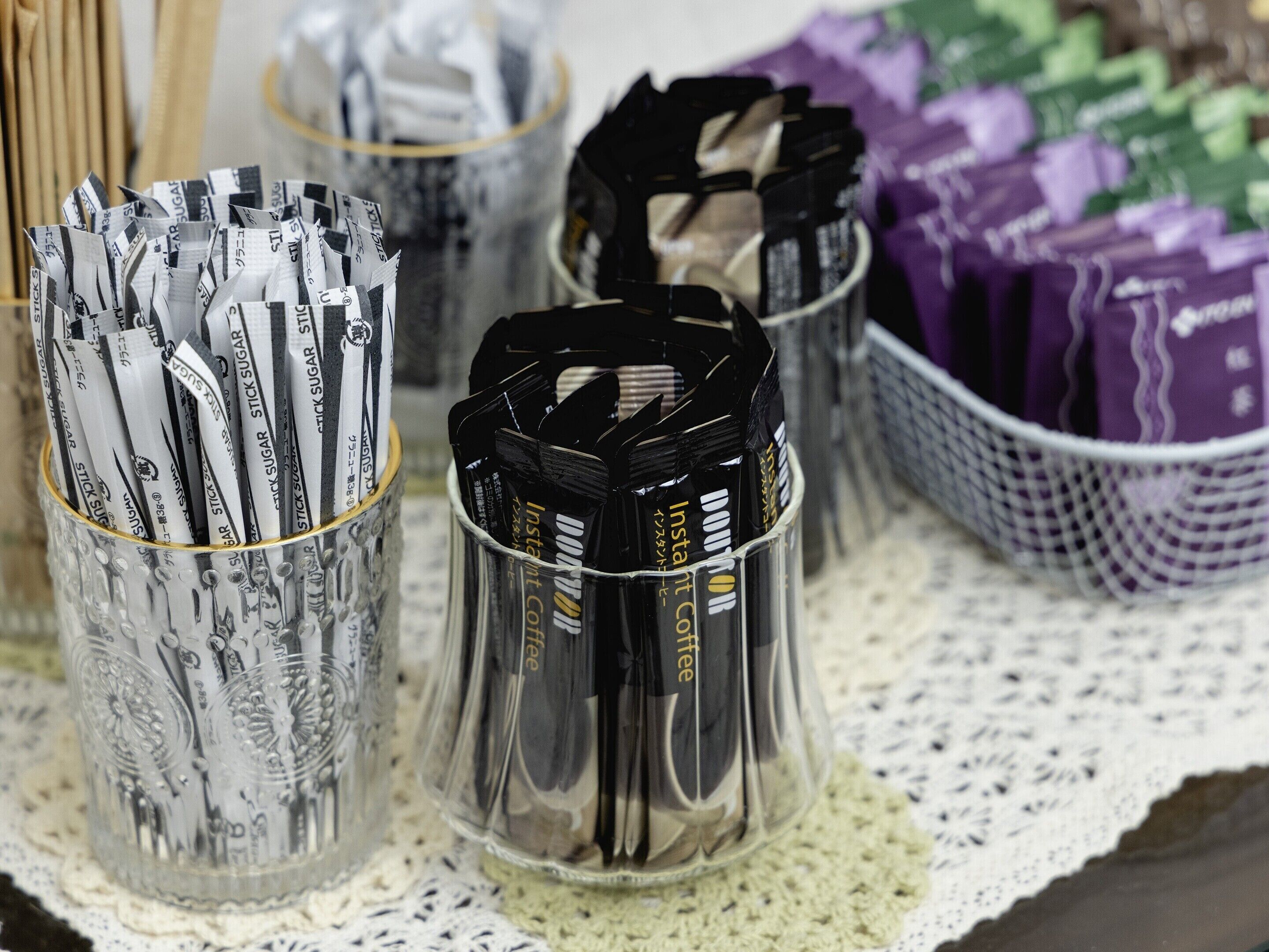 Des verres en verre transparent, remplis de sachets de café et de sachets de sucre, sont posés sur une nappe en dentelle.