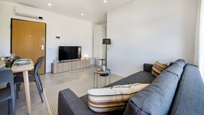 A modern living room with a grey sofa, a dining table with chairs, a flat-screen TV, and a small side table. - Perfect getaway with balcony and pool (Cabanas de tavira)