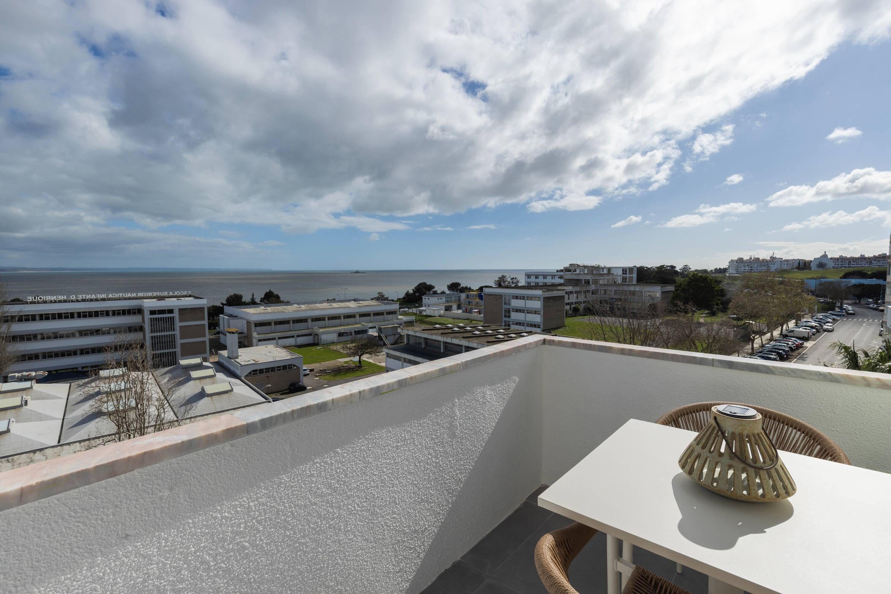 A balcony with a view of the sea and buildings, a table with a lamp, and a wicker chair.