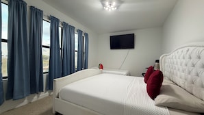 Ein Schlafzimmer mit einem großen Bett, einem flachbildfernseher an der Wand und Blick ins Freie durch das Fenster.