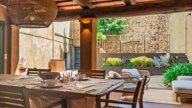 An outdoor dining area with a rustic stone wall, wooden furniture, and a hanging light fixture.