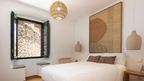 A bedroom with a large bed, a window with a view of a stone wall, a hanging light fixture, and a large abstract painting on the wall.