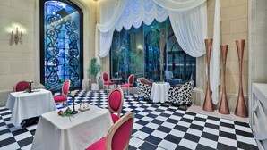A dining area with checkered flooring, tables with white tablecloths, red chairs, and a large window with draped curtains.