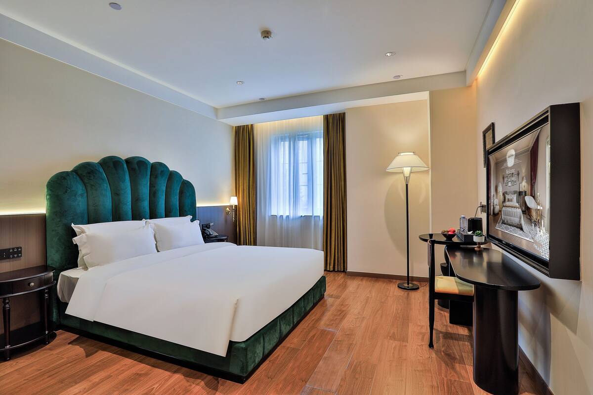 A hotel room with a large bed, a desk, a chair, a lamp, and a TV.
