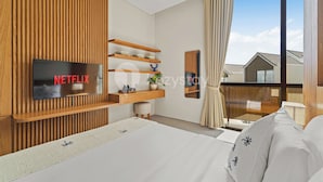 A modern hotel room with a large bed, a TV mounted on a wooden wall, a desk with a plant, and a view of the outside through a large window. - Manasa Villa by Kozystay - Jimbaran (Jimbaran)