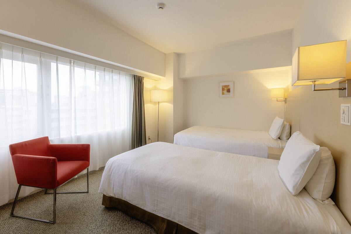 Ein Hotelzimmer mit zwei Betten, einem roten Sessel, einem großen Fenster mit Vorhängen und einer Stehlampe.