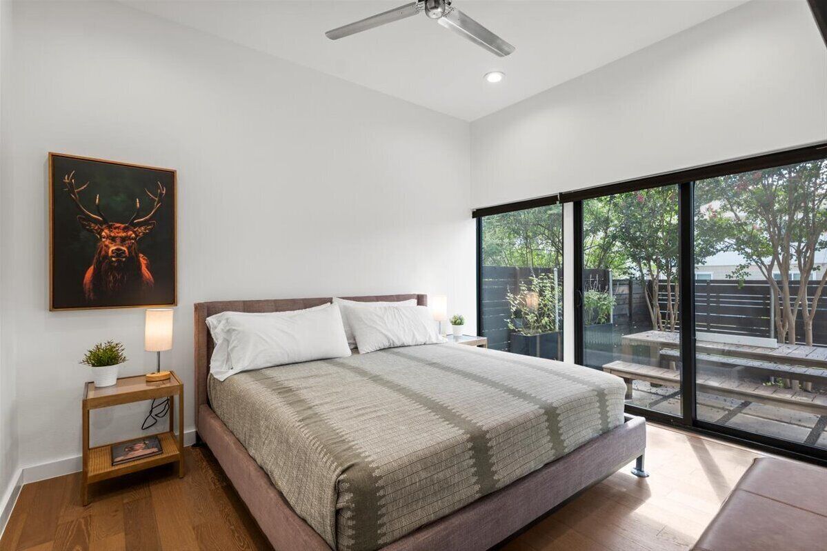 A modern bedroom with a large bed, a ceiling fan, a window with a view of the outdoors, and a framed deer artwork on the wall.