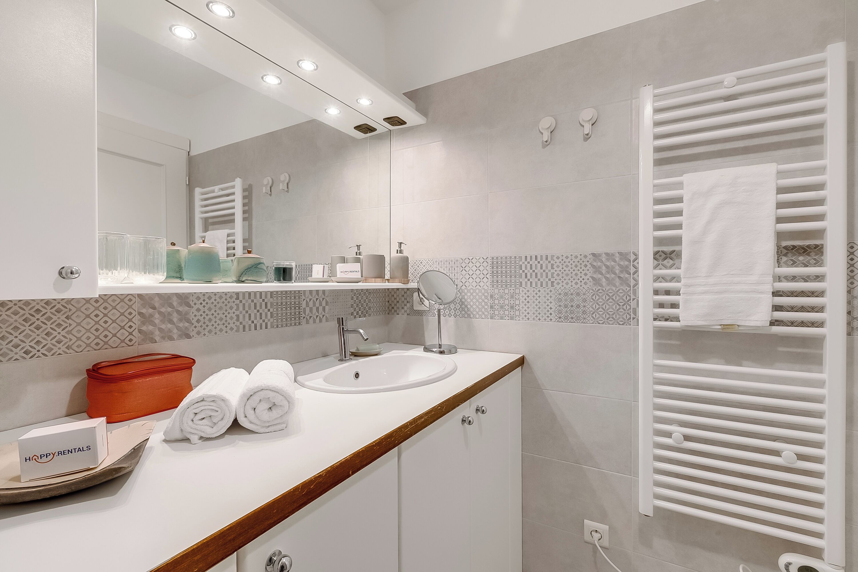 A modern bathroom with a white sink, mirror, and towel rack. There are toiletries on the counter and a sign that reads "HAPPY RENTALS".