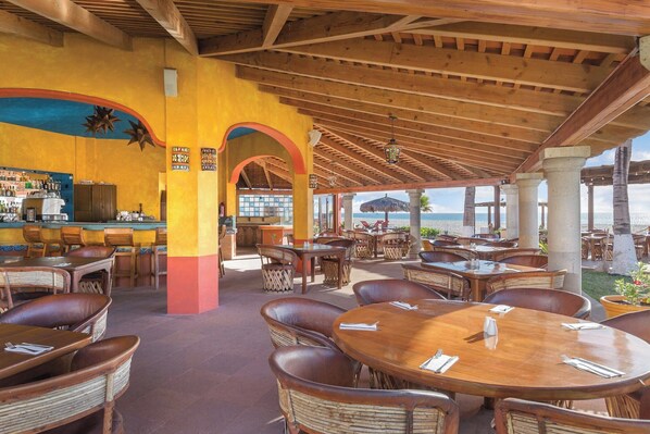 An outdoor restaurant with wooden tables and chairs, a bar area, and a view of the sea. - Cabo Oceanfront Resort Condos (San José del Cabo)