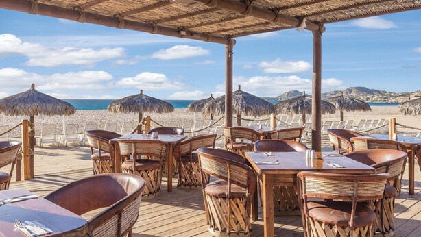 Un restaurant situé en bord de mer, avec des tables et des chaises en bois, des parasols en chaume et une vue sur la plage et l’océan.