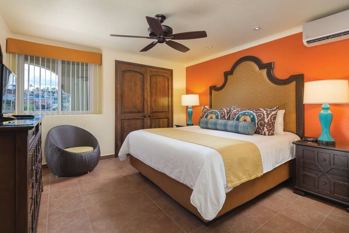 Une chambre d’hôtel comprenant un lit, un ventilateur de plafond, une télévision, une fenêtre avec des stores, une porte et une table de chevet avec une lampe.