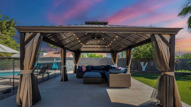 Un espacio de estar al aire libre cubierto con sofá y sillones, una piscina al fondo y un cielo al atardecer.