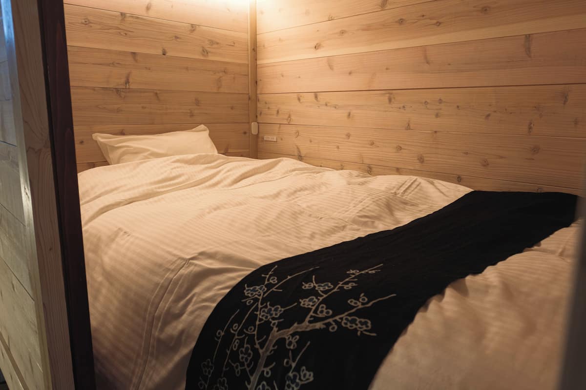 Une pièce aux murs lambrissés, avec un lit, du linge de lit blanc et une couverture noire ornée de broderies florales.