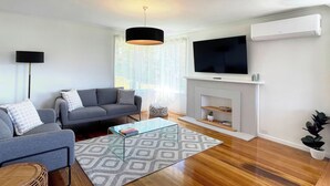 A modern living room with a grey sofa, a glass coffee table, a black pendant light, a flat-screen TV mounted above a fireplace, and a potted plant. - Quarry House - Bright and Renovated, near Hobart (Mornington)