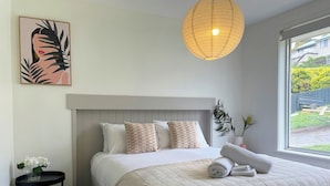 A neatly made bed with a quilted blanket and pillows, a bedside table with a vase of flowers, and a large window with a view of greenery outside. - Quarry House - Bright and Renovated, near Hobart (Mornington)