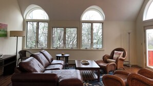 Ein geräumiges Wohnzimmer mit einer Ledersofa, einem Couchtisch und Korbstühlen. Zwei Deckenventilatoren und große Fenster sorgen für natürliches Licht.