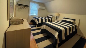 Ein kleines Zimmer mit zwei Betten, gestreifter Bettwäsche, einem Nachttisch, einer Lampe und einem Fenster mit Jalousien.
