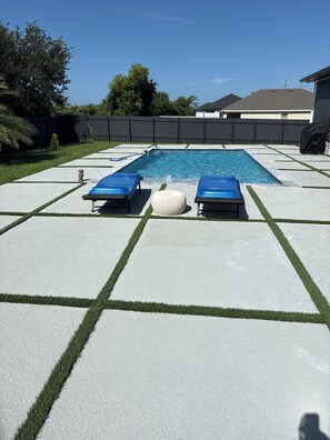 Ein rechteckiger Pool mit zwei blauen Liegestühlen und einem weißen Hocker auf einer Betonplattform, umrandet von grünem Rasen.