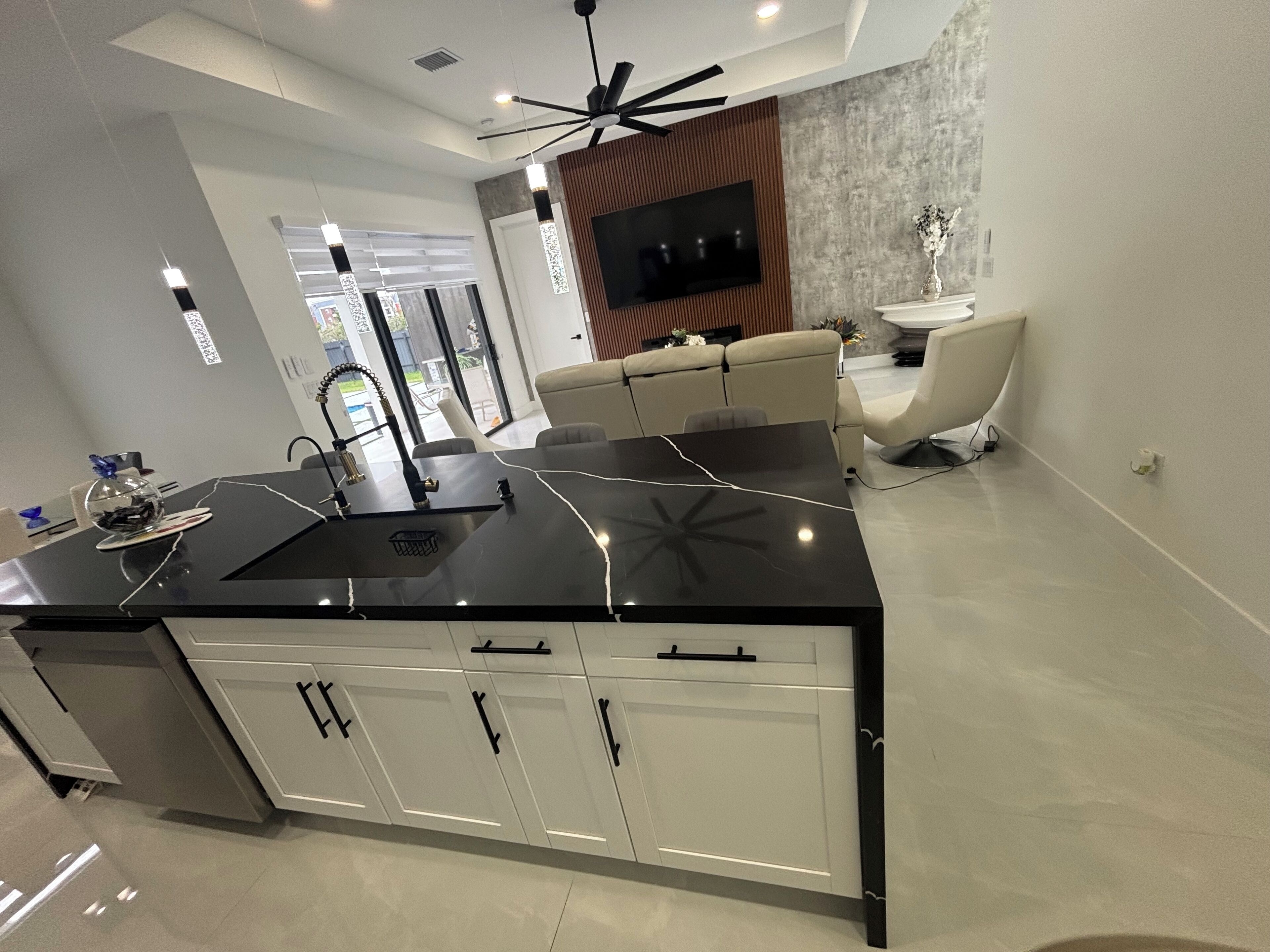 A modern kitchen with a black countertop, white cabinets, and a built-in oven. There is a dining area with a sofa and a TV mounted on the wall.