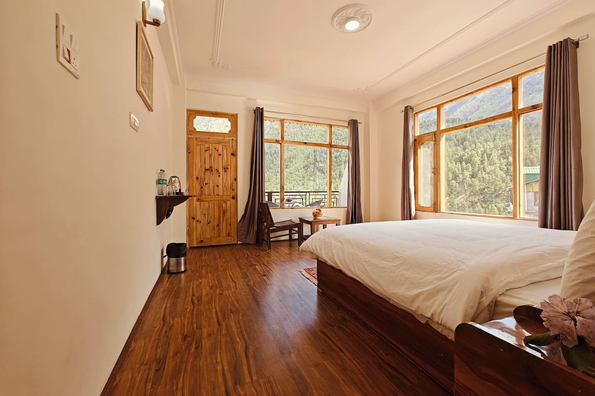 Quarto com piso de madeira, cama, cadeira, mesa, janela com vista para árvores e porta.