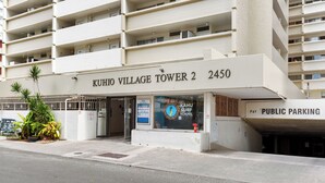 Un bâtiment portant l’inscription « KUHIO VILLAGE TOWER 2 2450 », avec une enseigne indiquant « KAHU SURF SCHOOL » et un panneau « PARCING PUBLIC ».