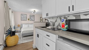Une cuisine moderne avec des meubles blancs, un four à micro-ondes et un évier. On y trouve un lit avec une tête de lit grise et une petite table de chevet avec une lampe.