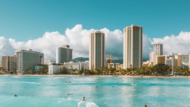 Un paysage urbain côtier avec de hauts immeubles et une plage où l’on peut voir des gens nager et faire de la planche à voile.