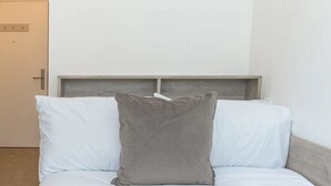 Ein Bett mit weißer Bettwäsche und einem grauen Kopfkissen, ein Holzkopfbrett und eine schlichte Wand.