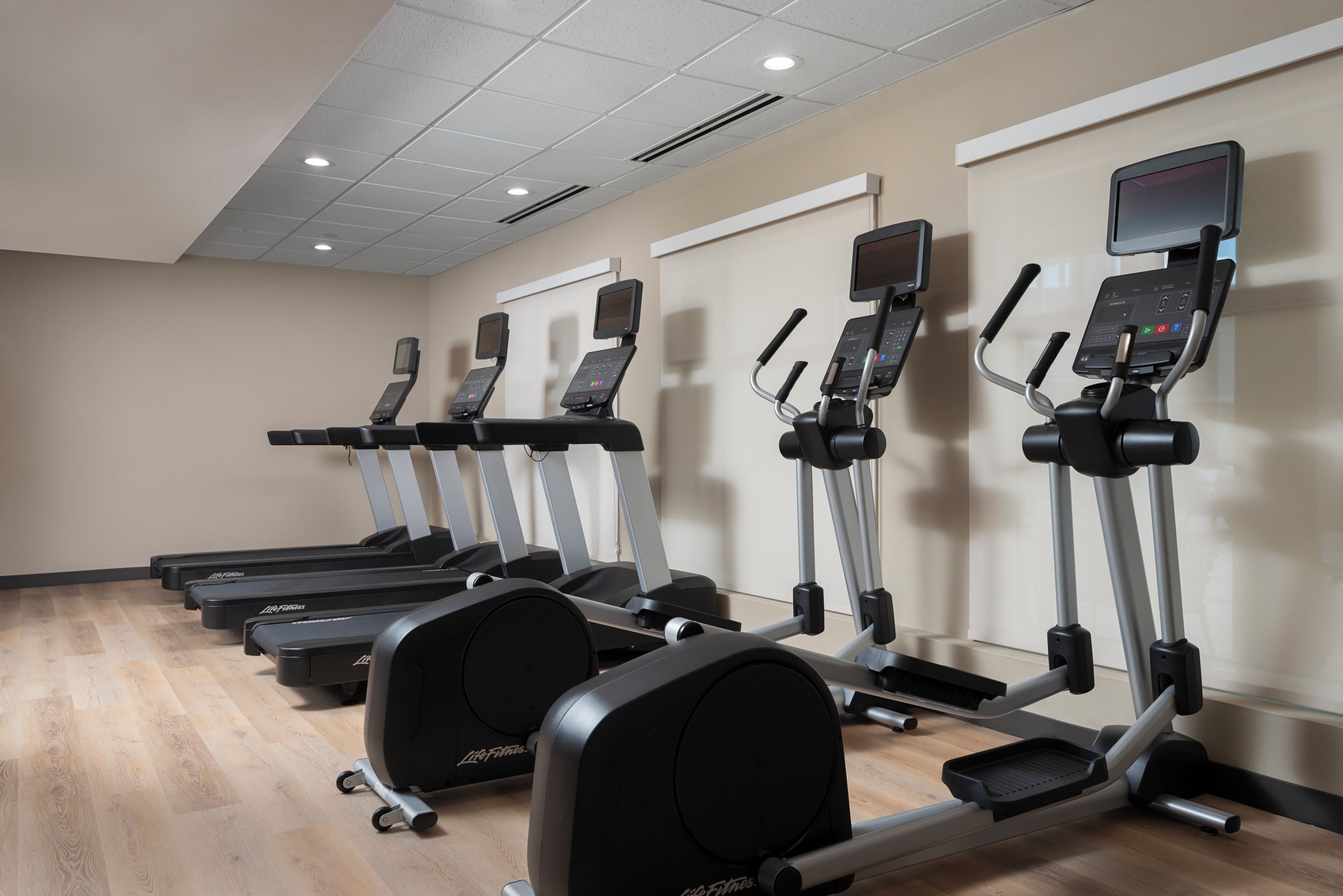 A row of treadmills and elliptical machines in a gym with a wooden floor and white walls.