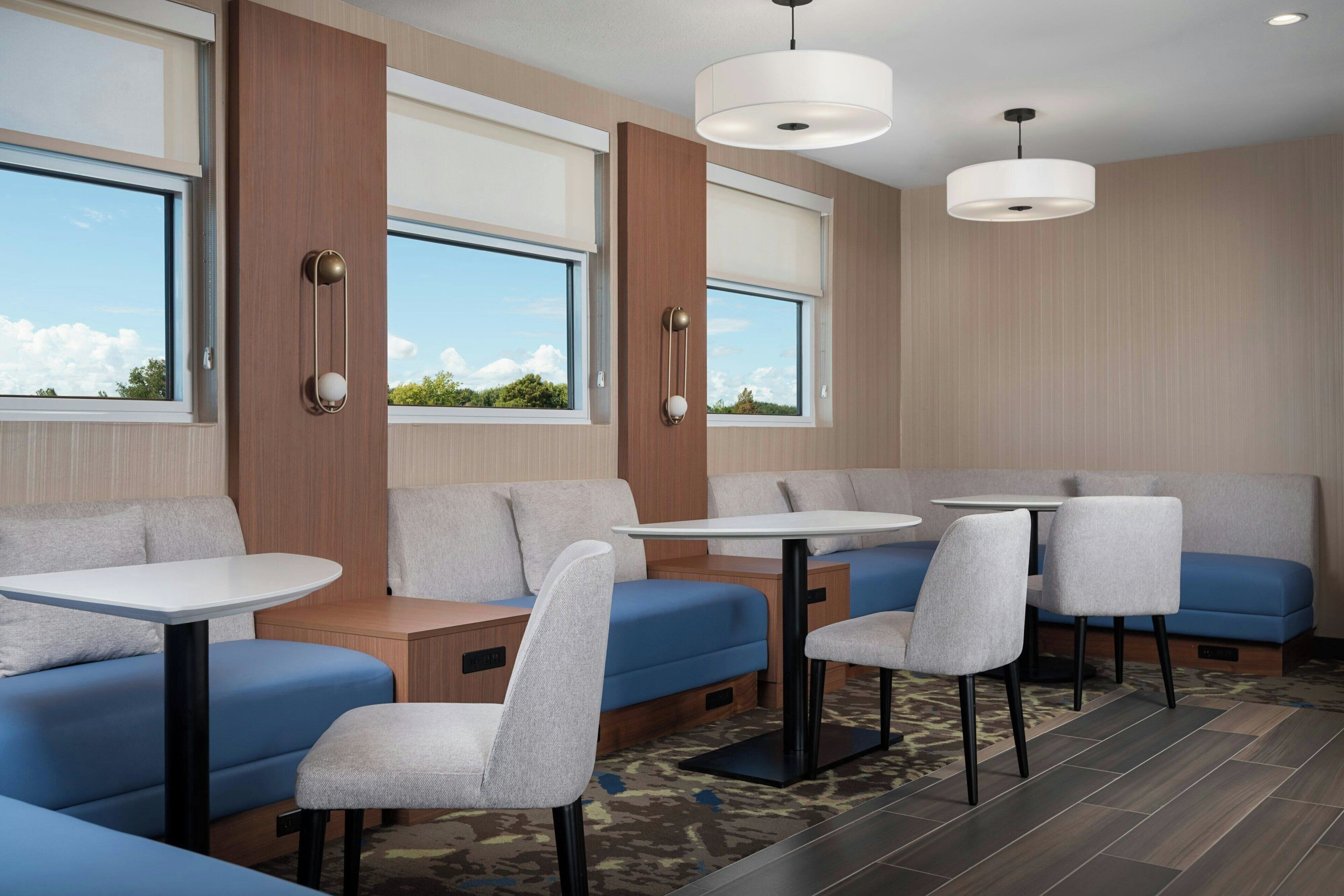 A modern hotel room with a seating area featuring blue sofas and white tables, large windows with blinds, and a ceiling-mounted light fixture.