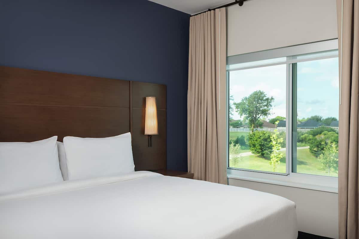 Ein Hotelzimmer mit einem großen Bett, einem Kopfteil mit einer Leuchte, einem Fenster mit Vorhängen und Blick auf Grünfläche.