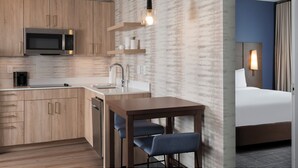 Habitación de hotel compacta con kitchenette, una cama, un escritorio con dos sillas y una lámpara de pared.