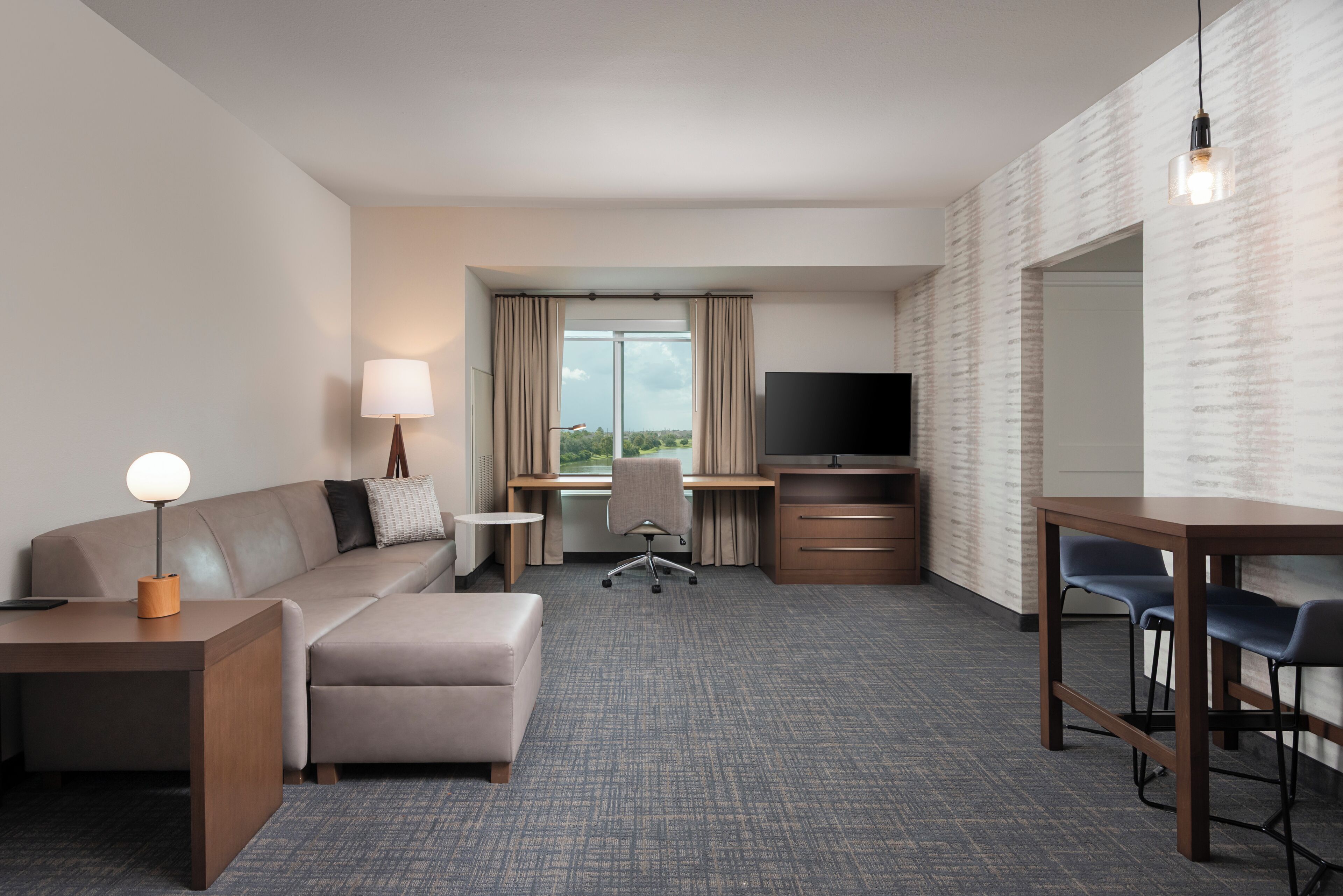 A hotel room with a sofa, ottoman, desk, TV, and a view of the outdoors.
