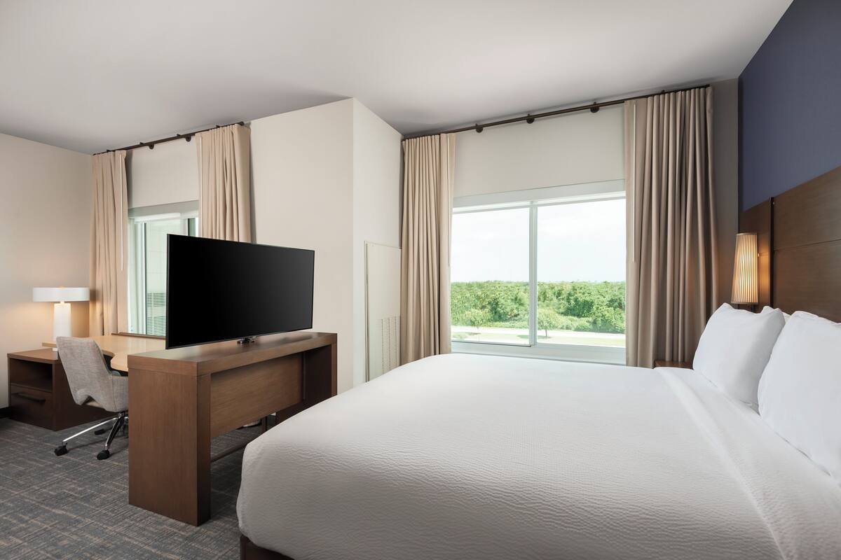 Ein Hotelzimmer mit einem großen Bett, einem Schreibtisch mit Fernseher, einem Sessel und einem Fenster mit Vorhängen.