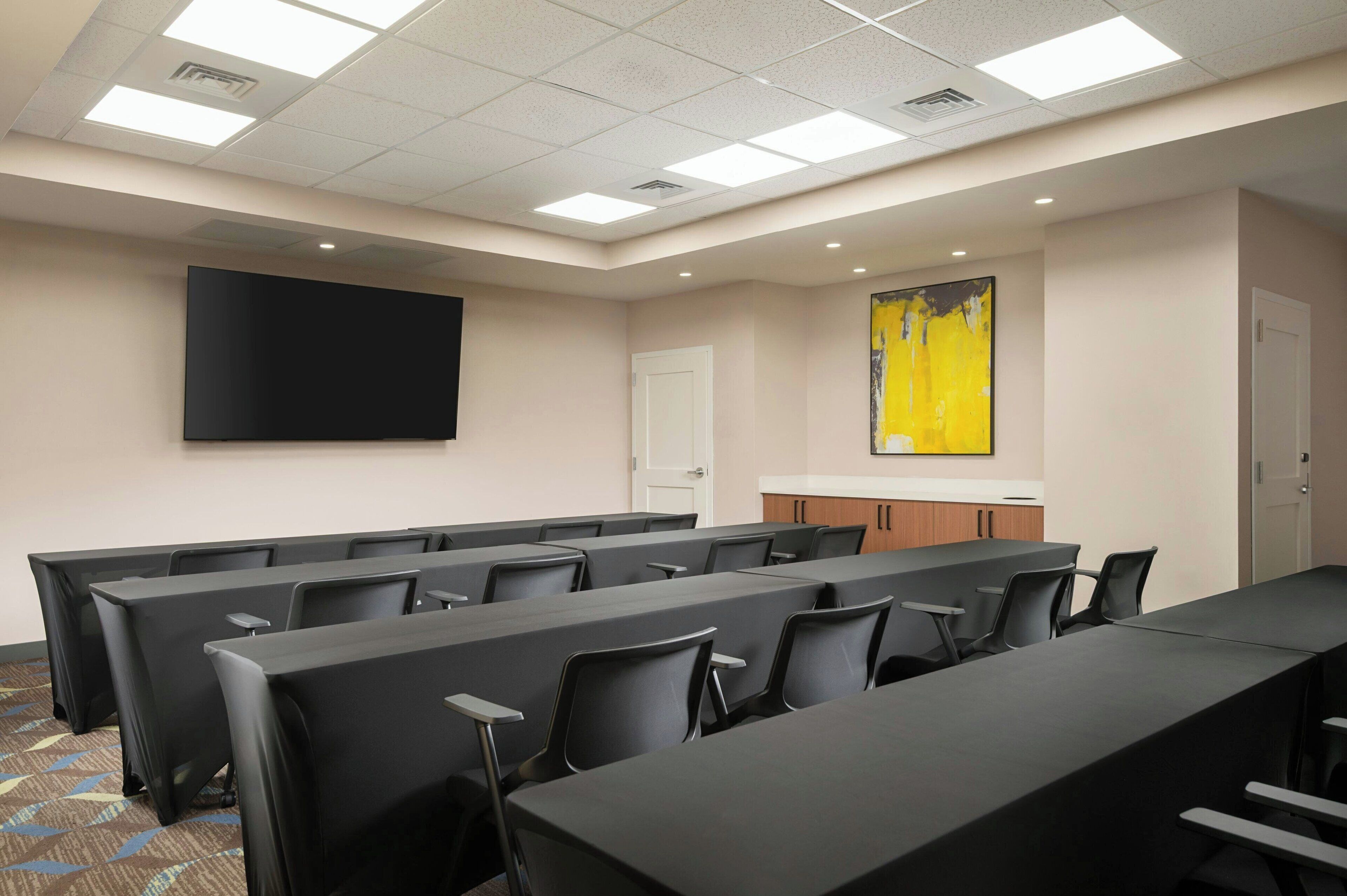 A conference room with rows of tables and chairs, a large flat-screen TV, a door, and a yellow abstract painting on the wall.