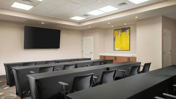 A conference room with rows of tables and chairs, a large flat-screen TV, a door, and a yellow abstract painting on the wall.