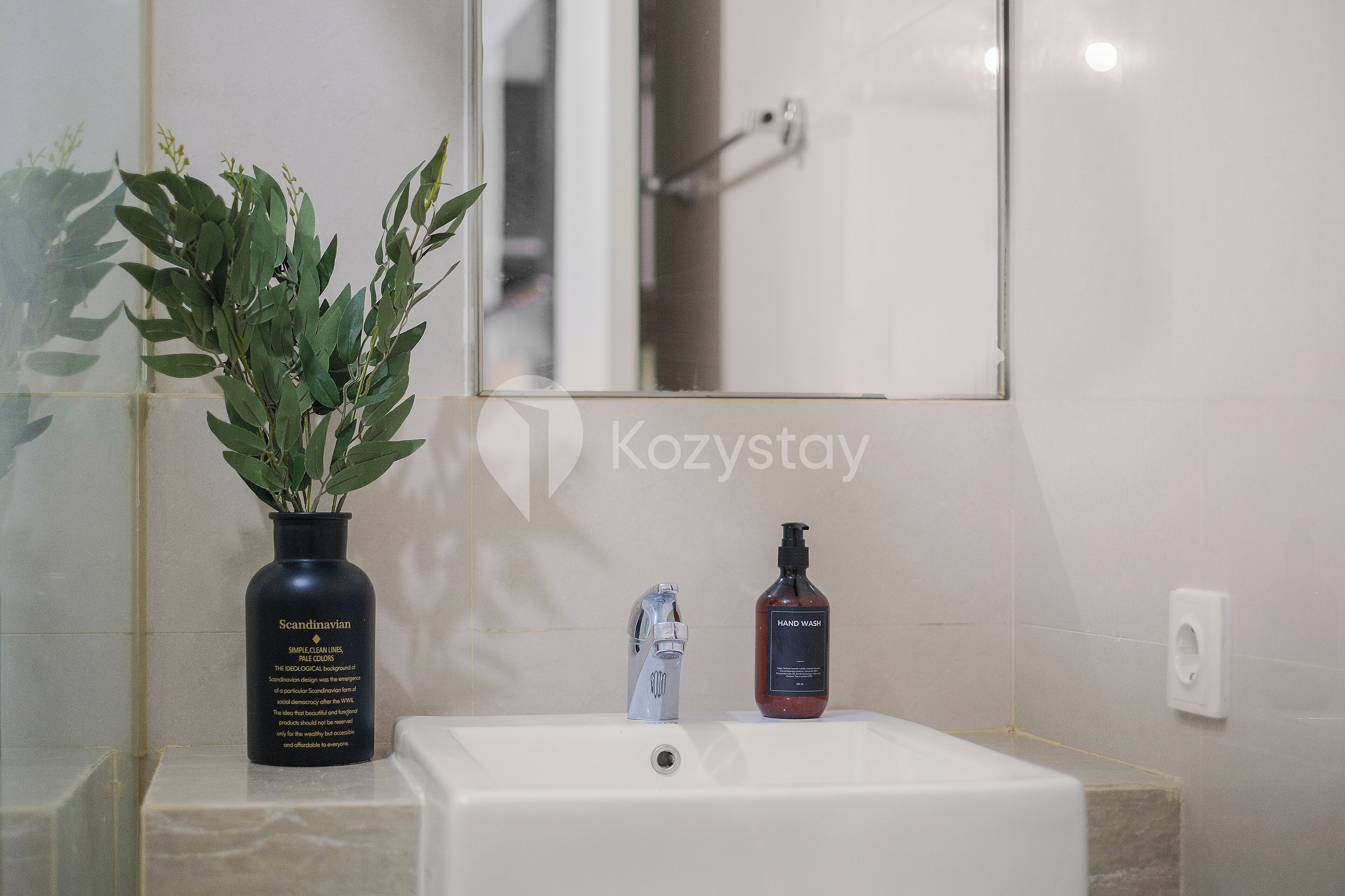 Un baño con lavamanos blanco, grifo plateado, botella negra de jabón de manos y una planta en maceta.