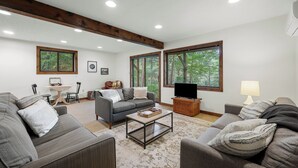 Amplia sala de estar con dos sofás, una mesa de centro y un mueble para televisión. Amplias ventanas ofrecen luz natural y vistas a árboles.