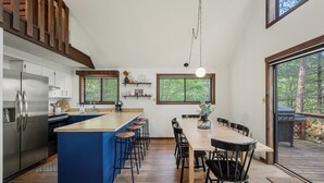 Una cocina moderna con una isla central, taburetes de bar y una mesa de comedor con sillas. Amplias ventanas que ofrecen luz natural y vistas al exterior.