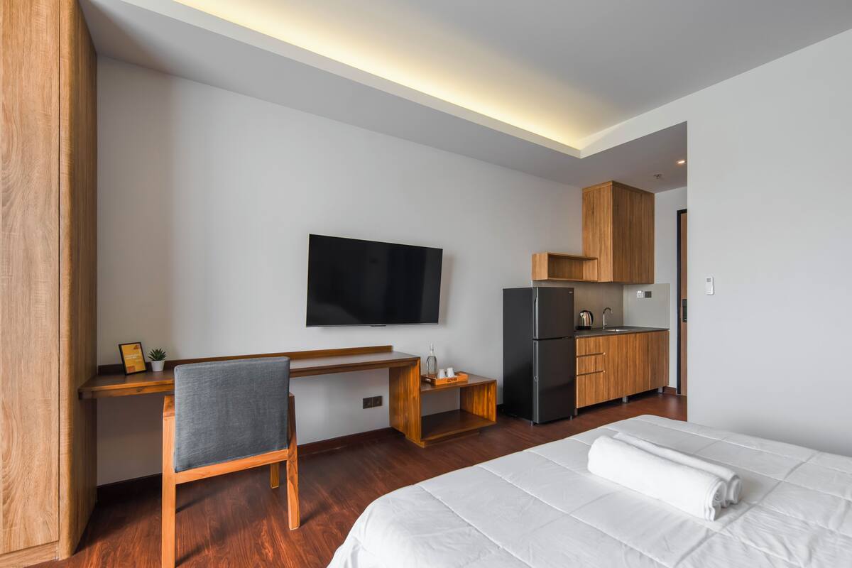 Ein modernes Hotelzimmer mit Schreibtisch aus Holz, einem Flachbildfernseher, einem Bett mit weißer Bettwäsche und einer Küchenzeile.