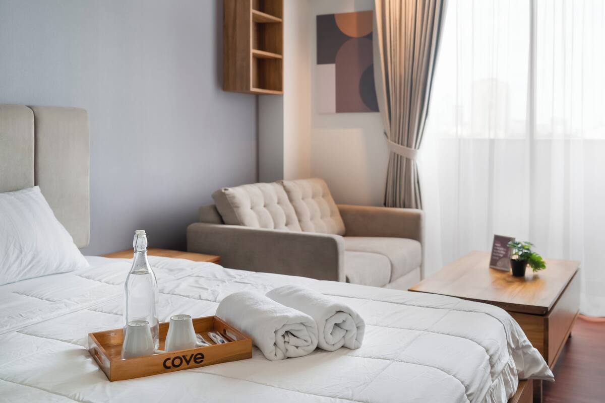 Ein Hotelzimmer mit einem ordentlich bezogenen Bett, einem Holftablett mit Toilettenartikeln, einem Sofa und einem kleinen Tisch mit einer Pflanze.