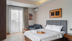 A modern hotel room with a large bed, a grey sofa, a wooden nightstand, and a bedside table. - Cove Amartha (Jakarta)
