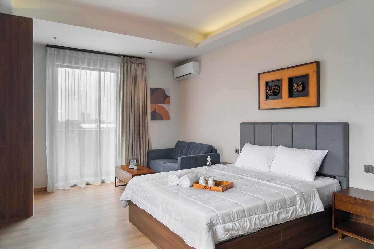 Ein modernes Hotelzimmer mit einem großen Bett, einem grauen Sofa, einem Holfnachtkästchen und einem Nachttisch.