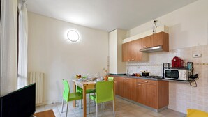 Une cuisine compacte avec des meubles en bois, un four à micro-ondes et un coin repas agrémenté de chaises vertes et d’une table dressée pour deux personnes.