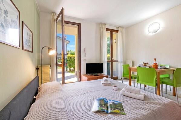 A hotel room with a bed, a TV, a dining table with chairs, and a view of the outdoors.